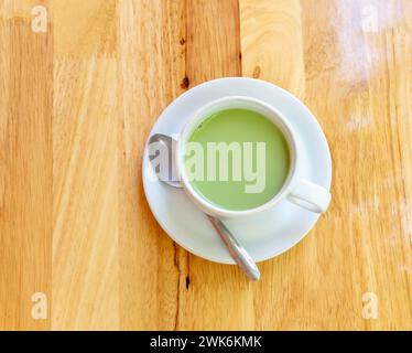 La vista dall'alto del tè verde matcha con latte caldo condensato viene servita in tazza bianca e in un piccolo piatto con un cucchiaio da tè su fondo marrone in legno. Foto Stock