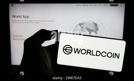 Persona che possiede un telefono cellulare con il logo del progetto di criptovaluta Worldcoin (Tools for Humanity) davanti alla pagina web. Mettere a fuoco il display del telefono. Foto Stock