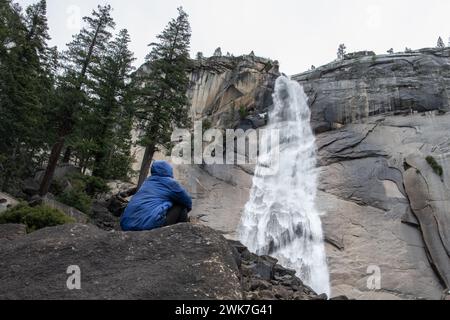 Un escursionista si siede di fronte e guarda in alto le acque che cadono nella cascata delle montagne della Sierra Nevada del Parco Nazionale di Yosemite, California, Stati Uniti. Foto Stock