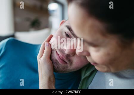 Ritratto di un giovane con la sindrome di Down con la madre a casa, abbracciando una guancia accarezzata. Concetto di amore e genitorialità bambino disabile. Foto Stock