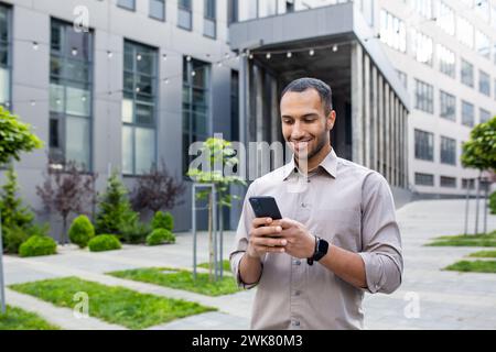 Sorridente giovane studente ispanico che usa il telefono, digita messaggi, controlla e legge notizie, cammina fuori dal campus universitario. Foto Stock