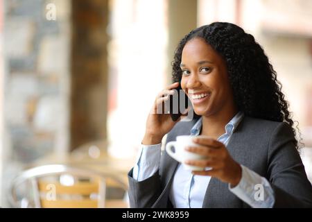 Un dirigente nero felice che beve e parla al telefono guardando voi nella terrazza di un bar Foto Stock