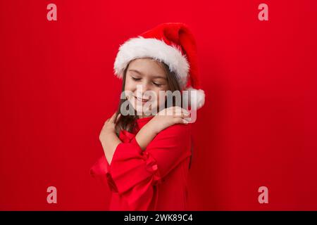 Adorabile ragazza ispanica, tutti sorridono, abbracciarsi in amore, irradiare felicità su un muro rosso isolato. indossato con un cappello di natale, esultante Foto Stock