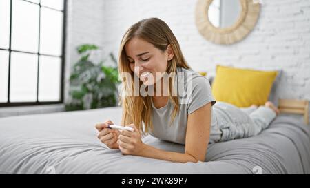 Giovane donna bionda che sorride gioiosamente, tiene un test di gravidanza positivo nella sua comoda camera da letto, si sente rilassata e felice sdraiata sul letto Foto Stock