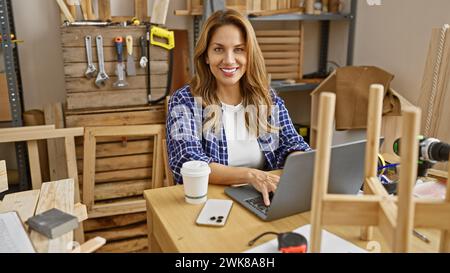 Una donna sorridente lavora con un notebook in un'officina di falegnameria circondata da utensili e legno. Foto Stock
