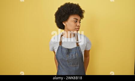 Ritratto di una donna afroamericana premurosa con capelli ricci su sfondo giallo, con grembiule in denim. Foto Stock