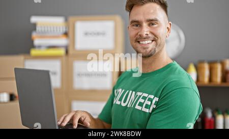 Uomo sorridente con la barba in una maglietta volontaria verde che utilizza un laptop in un magazzino con scatole sullo sfondo. Foto Stock