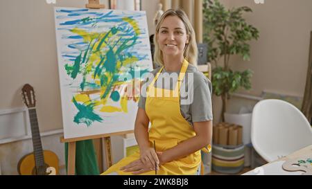 Donna sorridente in grembiule giallo posa in uno studio d'arte con pittura astratta e chitarra in sottofondo Foto Stock