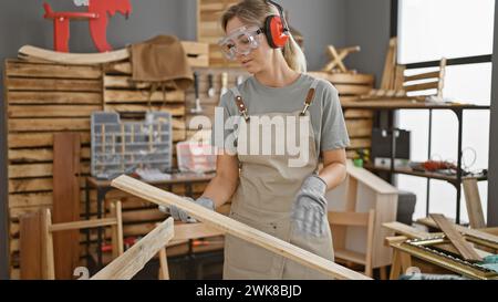 Una donna concentrata negli occhiali di sicurezza esamina la tavola in legno in un'officina di falegnameria ben attrezzata. Foto Stock