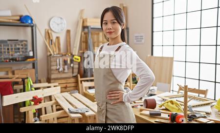 Una giovane donna asiatica sicura di sé che posa in un laboratorio di falegnameria ben attrezzato, traboccante di utensili e legname. Foto Stock