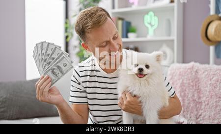 L'affascinante giovane uomo caucasico conta brillantemente le banconote in dollari americani seduto con il suo adorabile cane sul divano del soggiorno, irradiando la stalla Foto Stock