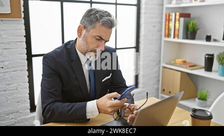 Giovane ispanico, lavoratore d'affari dai capelli grigi, che tiene in mano gli occhiali mentre invia SMS sullo smartphone negli stressanti interni dell'ufficio Foto Stock