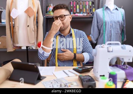 Uomo ispanico con barba disegnatore di sarte che lavora all'atelier odora qualcosa di puzzolente e disgustoso, intollerabile, trattenendo il respiro con le dita Foto Stock