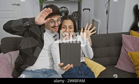 Una coppia allegra saluta durante una videochiamata nel loro accogliente salotto, condividendo un momento di connessione Foto Stock