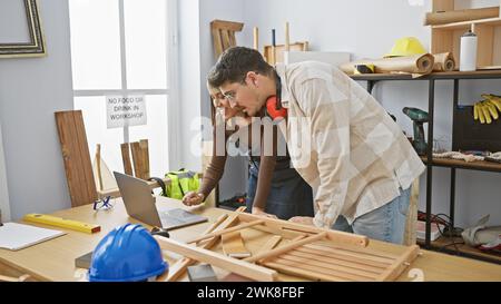 Un uomo e una donna collaborano in un'officina di lavorazione del legno, esaminando attentamente un telaio in legno vicino a vari utensili di falegnameria e attrezzature di sicurezza. Foto Stock