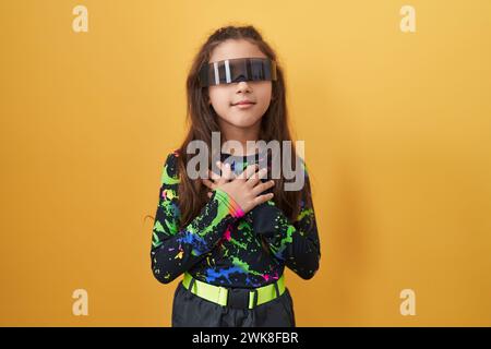 Adorabile ragazza ispanica, con le mani sul petto, sorridente e riconoscente con gli occhi chiusi che indossano occhiali per realtà virtuale. un'immagine di salute Foto Stock