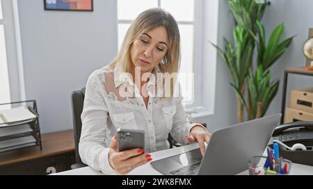 Una donna caucasica professionista utilizza uno smartphone e un laptop in un ufficio moderno, ritraendo una donna d'affari matura che lavora in multitasking. Foto Stock