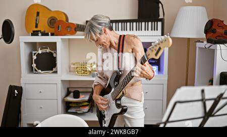 Una donna matura che suona una chitarra elettrica concentrata in uno studio di musica casalinga con vari strumenti. Foto Stock