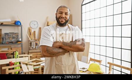 Uomo sorridente barbuto con le braccia incrociate in un laboratorio di falegnameria illuminato dal sole, con grembiule indicativo del lavoro. Foto Stock