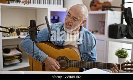 Un anziano ispanico con la barba canta mentre suona una chitarra acustica in uno studio domestico. Foto Stock