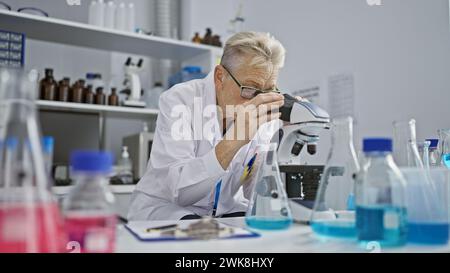 Scienziata senior con i capelli grigi dedicata immersa in un intenso lavoro di ricerca sul microscopio a livello di dottorato in laboratorio Foto Stock