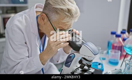 Scienziata senior con i capelli grigi dedicata immersa in un intenso lavoro di ricerca sul microscopio a livello di dottorato in laboratorio Foto Stock