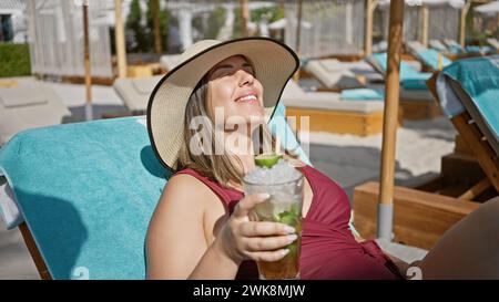 Una donna sorridente in un cappello da sole gode di un mojito mentre si rilassa su una sedia da spiaggia in un resort di lusso. Foto Stock