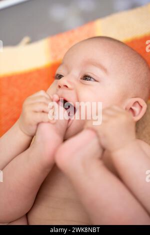 piccola bambina sorridente sdraiata su una coperta che le mette i piedi in bocca a causa del disagio causato dalla dentizione. concetto di apparizione dei primi denti Foto Stock