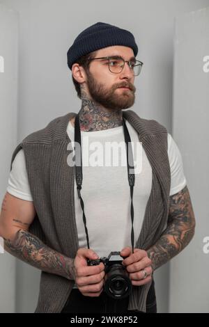 Brutale e affascinante fotografo professionista Hipster Man con occhiali con tatuaggi con cappello e maglione in maglia tiene Una fotocamera e si trova in piedi Foto Stock