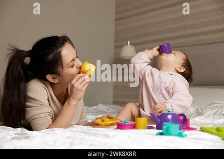 madre felice e figlia piccola che fingono di bere il tè da tazze di plastica giocattolo in camera da letto, la famiglia si diverte Foto Stock