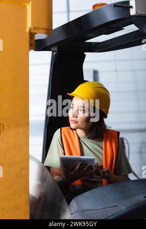 lavoratrice di magazzino pensiva con elmetto e giubbotto di sicurezza con tablet e seduta su un carrello elevatore Foto Stock