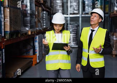 supervisor discussing productivity of warehouse with female employee in hard hat with clipboard Foto Stock