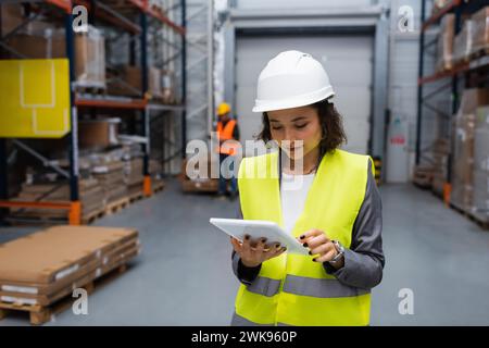 supervisore di magazzino donna con elmetto e giubbotto di sicurezza focalizzato sul tablet digitale durante il lavoro Foto Stock