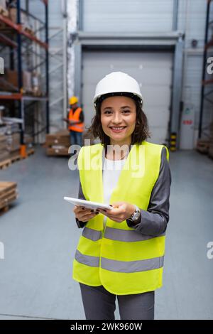 supervisore di magazzino donna felice con elmetto e giubbotto di sicurezza focalizzato sul tablet digitale durante il lavoro Foto Stock