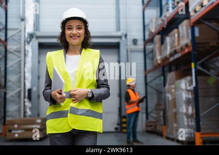 supervisore di magazzino donna gioiosa con elmetto e giubbotto di sicurezza focalizzato sul tablet digitale durante il lavoro Foto Stock