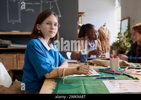 Vista laterale del ritratto di adolescente con protesi che disegnano a mano gli schizzi di abiti in classe sartoriale e guardano la fotocamera, copiano lo spazio Foto Stock