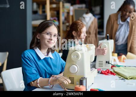 Vista laterale del ritratto di una ragazza con la mano protesica che usa la macchina da cucire in classe e guarda la fotocamera Foto Stock