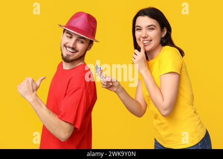 Giovane donna che attacca il clown di carta sulla schiena della sua amica su sfondo giallo. Festa del giorno dello scemo d'aprile Foto Stock