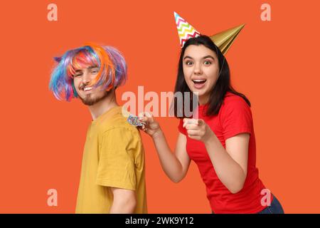 Giovane donna che attacca il pesce di carta sulla schiena della sua amica su sfondo arancione. Festa del giorno dello scemo d'aprile Foto Stock