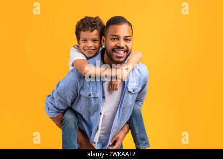 Padre e figlio neri felici che condividono un abbraccio commovente sullo sfondo giallo dello studio Foto Stock