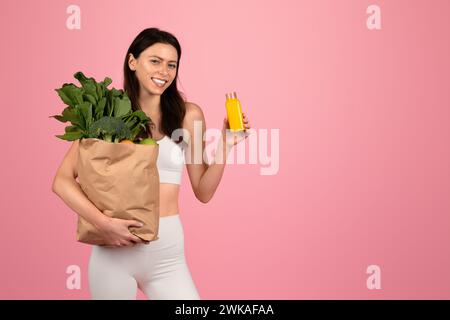 Donna in forma sorridente con abiti da allenamento bianchi che reggono un sacchetto di carta pieno di verde e una bottiglia di succo di frutta Foto Stock