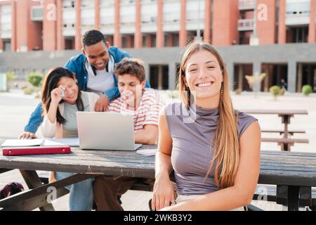 Giovane studentessa caucasica che sorride e guarda la macchina fotografica seduta con le sue compagne di classe nel campus universitario. Studentessa bionda che guarda davanti dietro i suoi amici multirazziali mentre usano un laptop. Foto di alta qualità Foto Stock