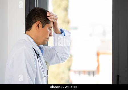 Shot di un giovane medico maschile che osserva stressato mentre si alza in piedi in una finestra in un ospedale Foto Stock