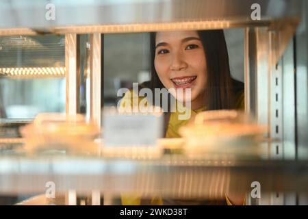 Proprietaria di una piccola impresa sorridente che organizza torte in vetrina nel suo negozio di panetteria Foto Stock