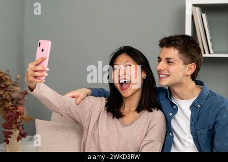 Uomo e donna allegri che ridono in una videochiamata con lo smartphone. Diverse giovani coppie sul divano nel loro salotto che si fanno un selfie. Foto Stock