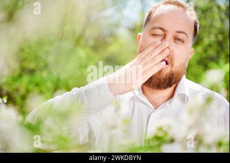 L'uomo allergico soffre di allergia stagionale in primavera nel giardino fiorito in primavera. Primo piano di un uomo barbuto che starnutisce di fronte all'albero in fiore. Concetto di allergia primaverile. Foto Stock