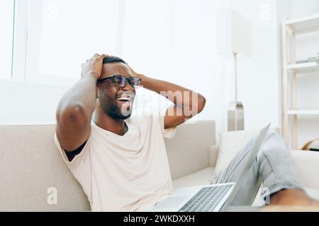 Freelance afroamericano sorridente che lavora su un computer portatile mentre si siede su un divano moderno in un accogliente soggiorno, questa immagine cattura un giovane e felice Foto Stock