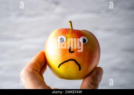 Augusta, Baviera, Germania - 18 febbraio 2024: Un uomo tiene in mano una mela dal volto triste *** Ein Mann hält einen Apfel mit einem traurigen Gesicht Foto Stock