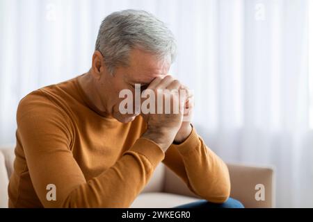 Triste uomo anziano seduto sul divano a casa, toccando la testa Foto Stock