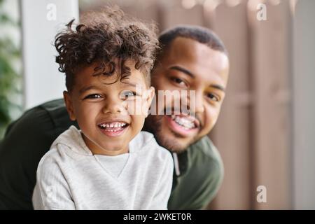 ritratto di famiglia di un uomo afroamericano allegro in bretelle che abbraccia il figlio riccio mentre guarda la macchina fotografica Foto Stock